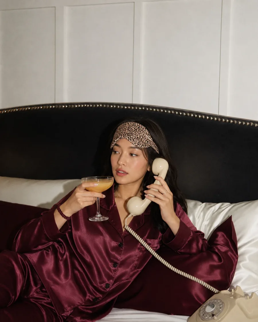 Woman in burgundy satin pajamas on a rotary phone in bed, holding a coupe glass